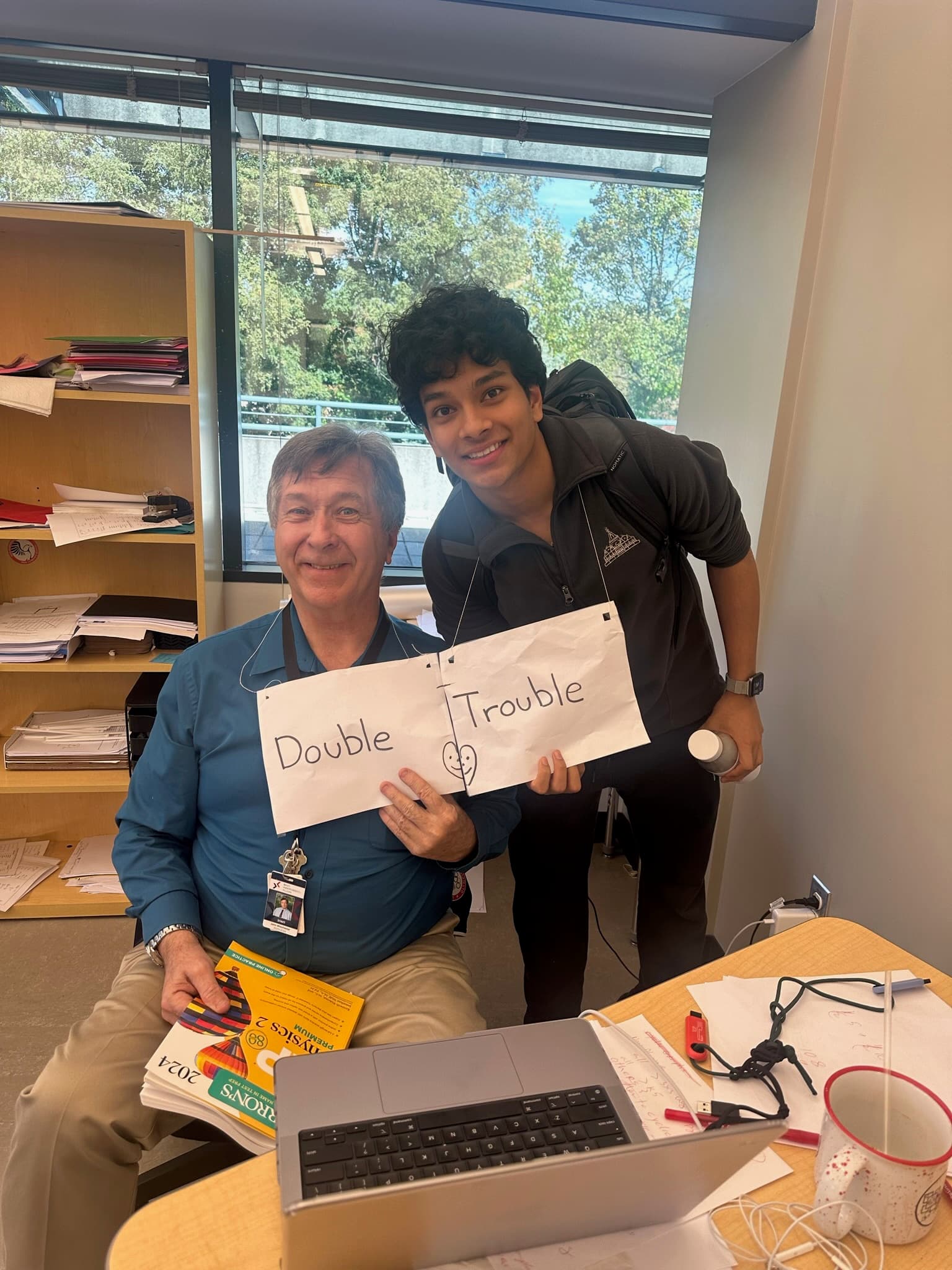 A teacher at a desk and a student behind him holding two paper signs reading Double and Trouble