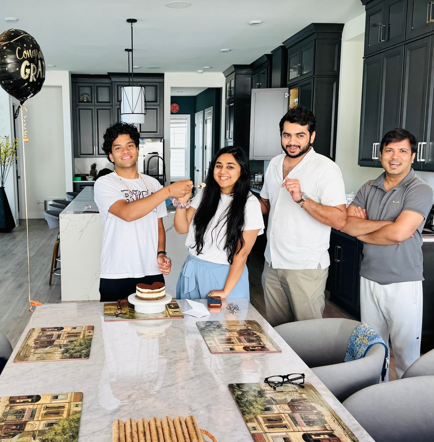 Four people around a kitchen island with a tiramisu cake on a stand, a Congrats Grad balloon at the left, one of them being fed the first bite