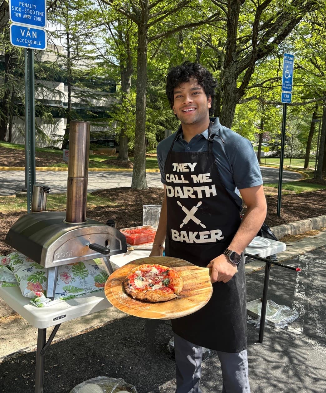A cook in a Darth Baker apron holding a margherita on a wooden peel beside an Ooni oven set up in a car park
