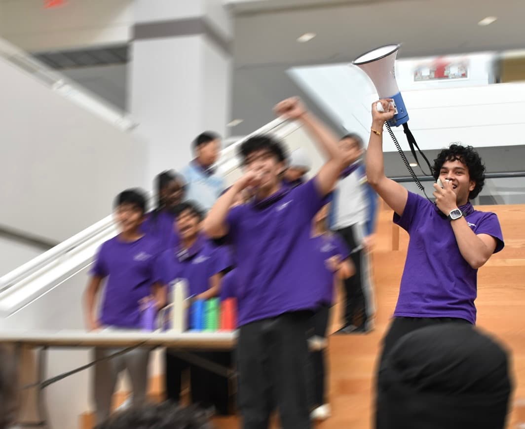 A student on a megaphone at the front of a school hall, a line of classmates in matching purple shirts cheering behind him