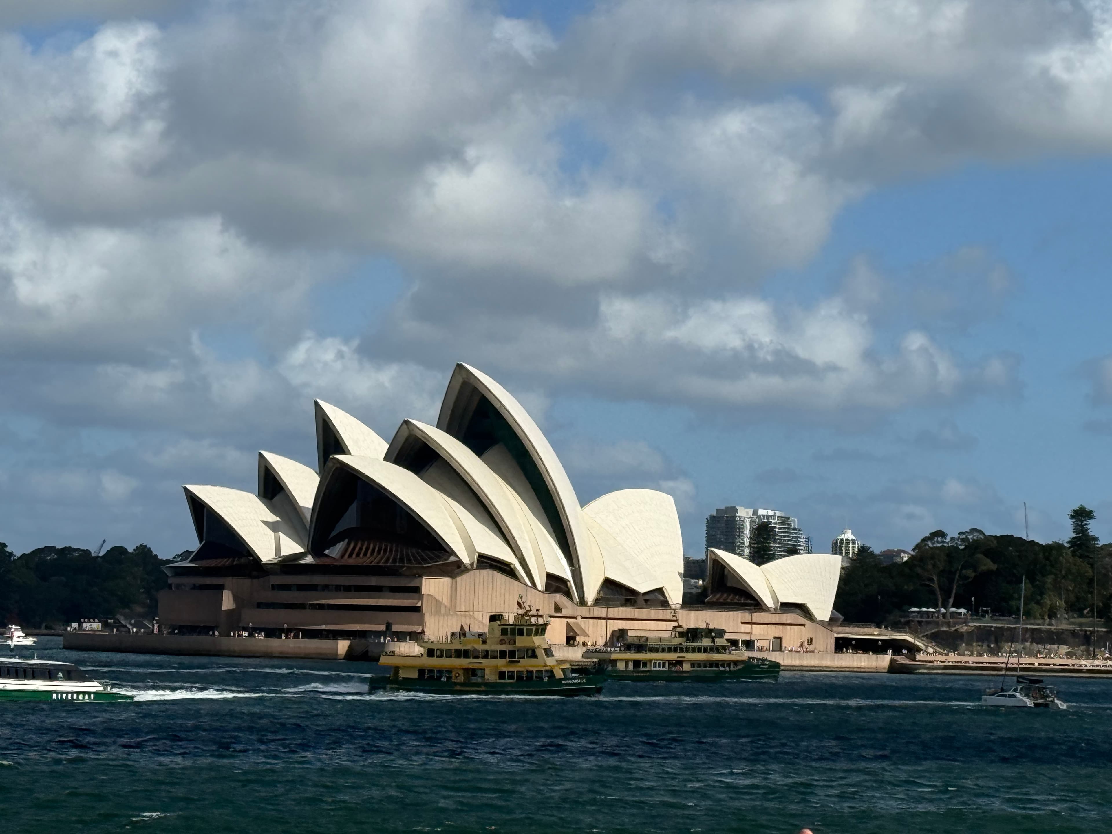 The Sydney Opera House seen across the harbour, two ferries passing in front of it under heavy cloud