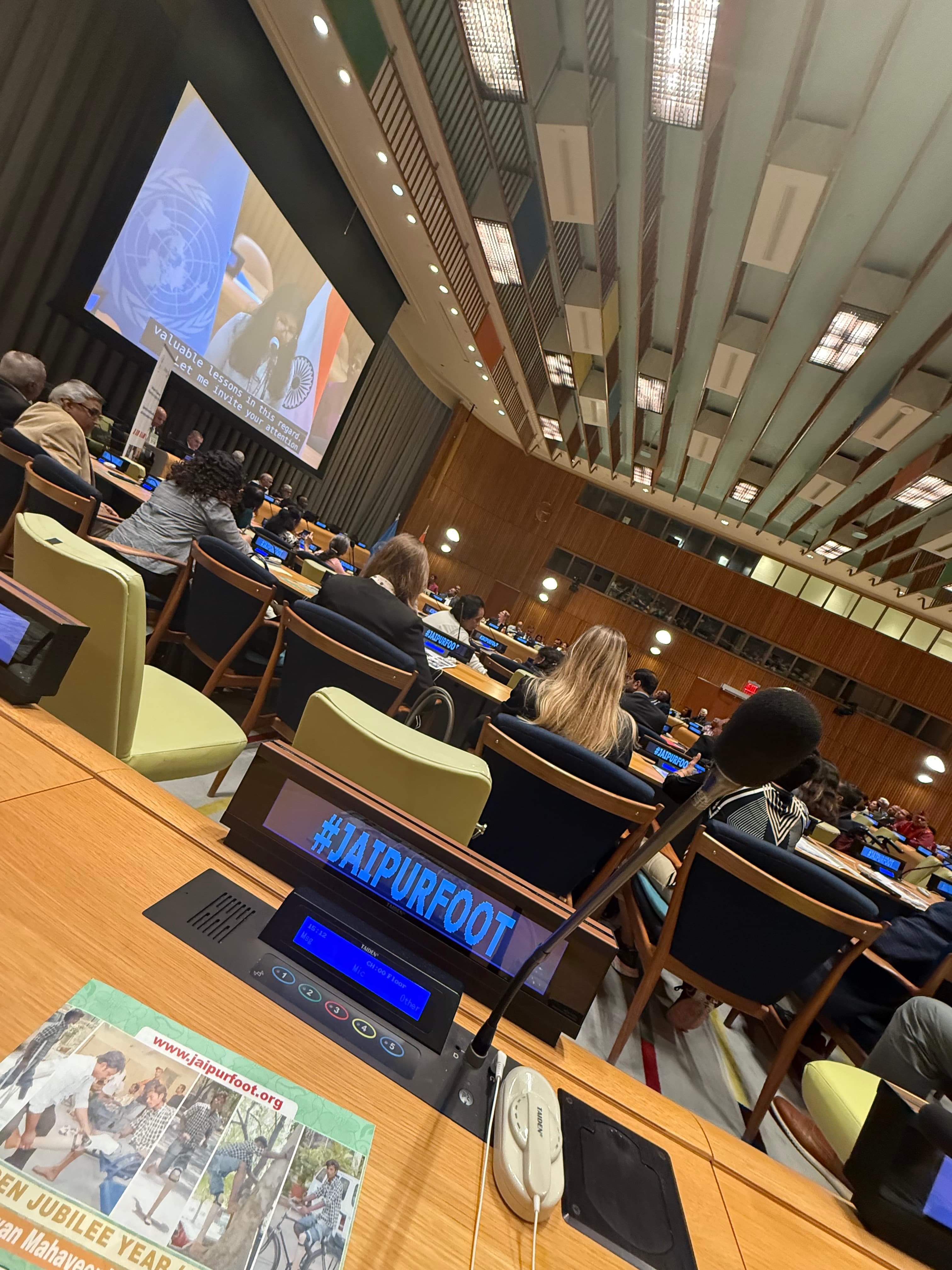 A United Nations chamber seen from a delegate's desk, screens along the benches reading JAIPURFOOT