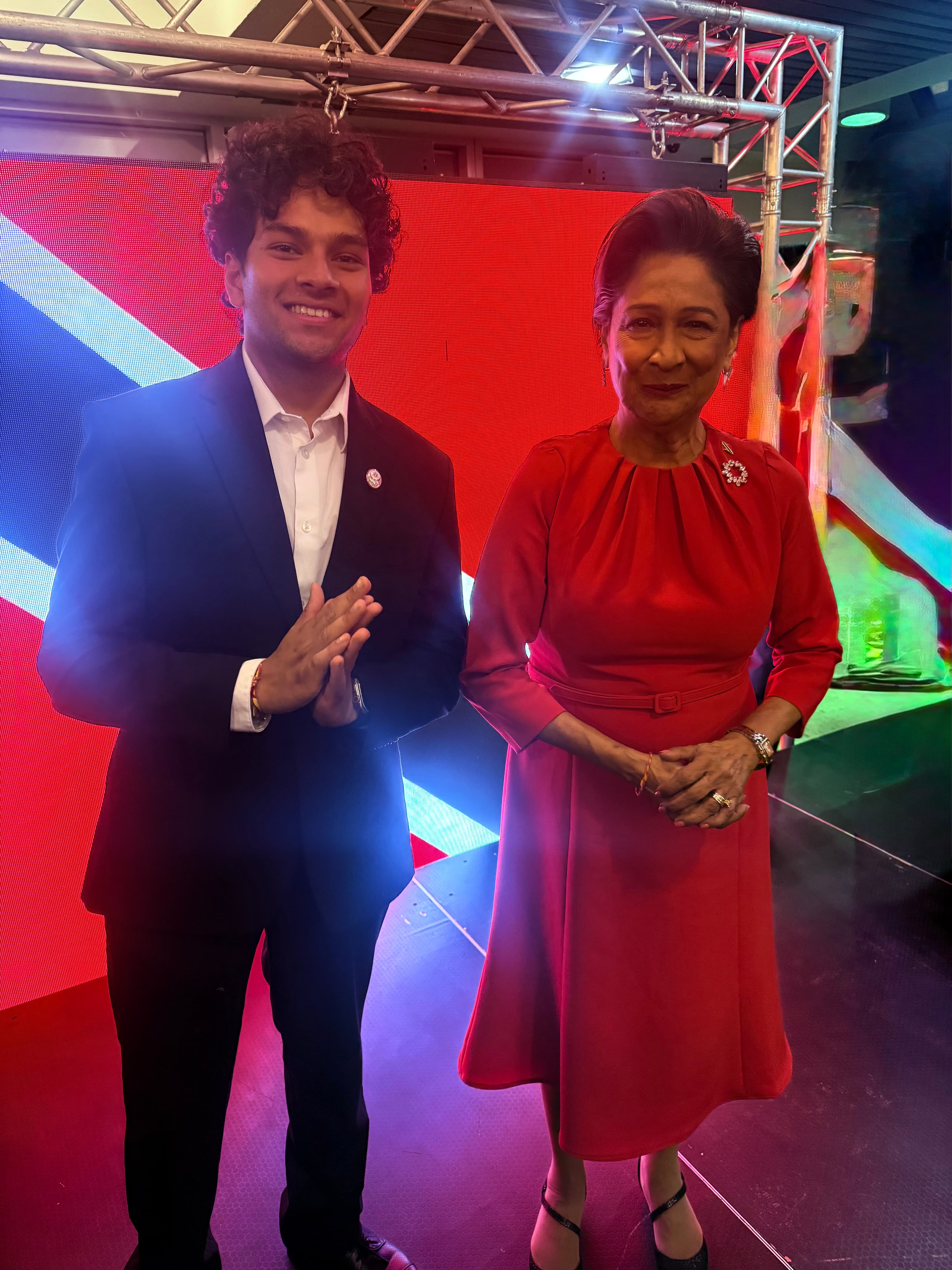 Two people standing together in front of a lit stage screen, one in a dark suit and one in a red dress
